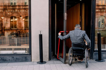 Rückansicht eines behinderten reifen Mannes, der auf einem Rollstuhl sitzt, während er in ein Geschäft geht - MASF13009