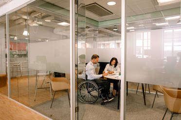 Businessman sitting on wheelchair while working with female colleague in board room at work place seen through doorway - MASF13019