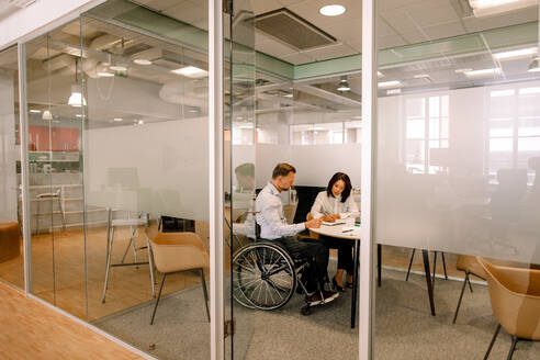 Geschäftsmann im Rollstuhl sitzend bei der Arbeit mit einer Kollegin im Sitzungssaal am Arbeitsplatz, gesehen durch die Türöffnung - MASF13019