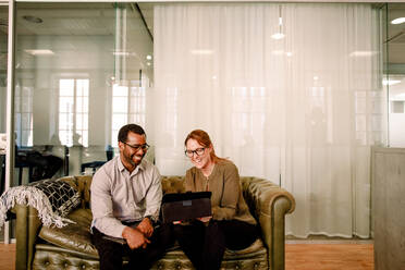 Fröhliche Geschäftskollegen schauen auf ein digitales Tablet, während sie auf dem Sofa im Büro sitzen - MASF13040