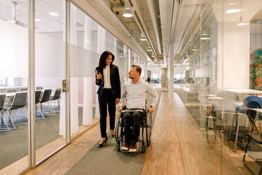Lächelnde Geschäftsfrau, die einen behinderten Kollegen im Rollstuhl sitzend im Korridor am Arbeitsplatz betrachtet - MASF13044