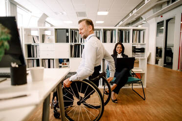 Ein behinderter Mann sitzt im Rollstuhl, während eine lächelnde Geschäftsfrau einen Laptop am Arbeitsplatz benutzt - MASF13050