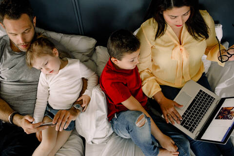 Directly above shot of parents using technologies while sitting with children on bed stock photo