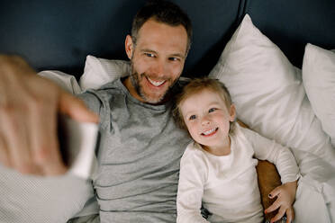 High angle view of happy father taking selfie with smiling daughter on bed in hotel - MASF13069