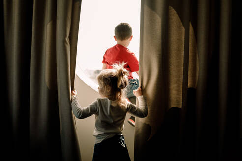 Rückansicht eines Mädchens, das einen Vorhang hält, während sein Bruder am Fenster eines Hotelzimmers sitzt - MASF13072