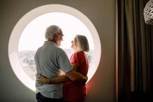 Senior couple embracing while standing by window in hotel room - MASF13083