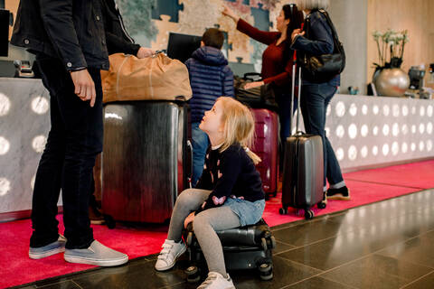 Mädchen sitzt auf einem Koffer und sieht den Vater an, während die Familie im Hintergrund steht, lizenzfreies Stockfoto