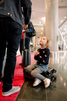 Mädchen in voller Länge mit verschränkten Händen, das seinen Vater anschaut, während es auf einem Koffer im Hotel sitzt - MASF13104