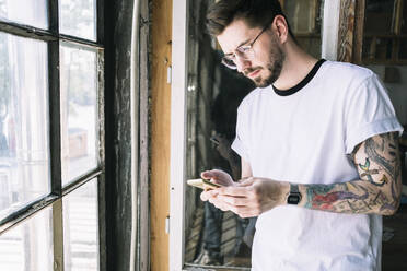 Confident male hipster hacker text messaging through smart phone by window at creative office - MASF13116