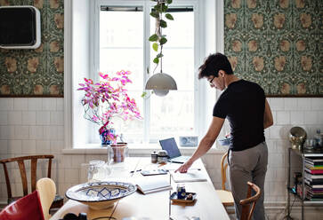 Rear view of young man reading book on table while standing at home - MASF13119