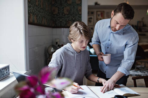 Father holding coffee cup while assisting son in studying at home - MASF13121