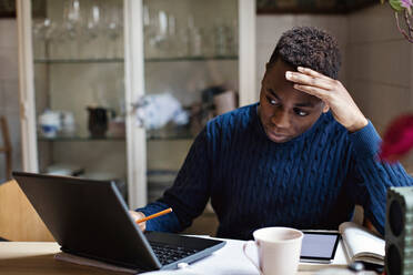 Worried teenage boy looking at laptop while doing homework on table - MASF13144