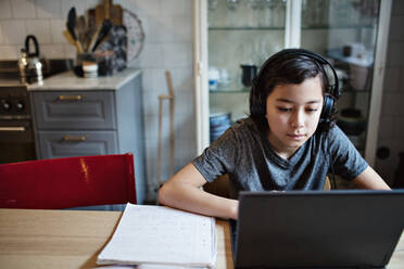 Boy wearing headphones while using laptop during homework at home - MASF13147