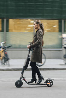 Full length side view of businesswoman riding electric push scooter on street in city - MASF13260