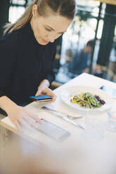 Selbstbewusste Unternehmerin, die ein Mobiltelefon benutzt, während sie mit einer Kreditkarte in einer Cafeteria im Büro sitzt - MASF13266