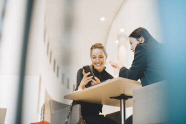 Low angle view of smiling businesswoman sharing smart phone with female coworker at table in cafeteria - MASF13274