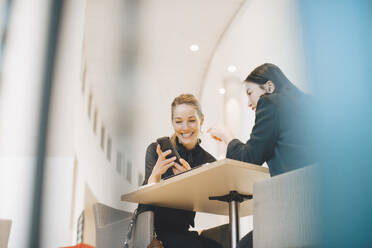 Low angle view of smiling businesswoman sharing smart phone with female colleague at table in office - MASF13276