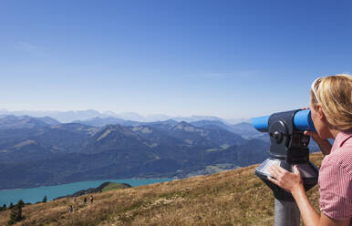 Frau betrachtet die Berge durch ein Münzfernglas vom Schafberg aus vor blauem Himmel - GWF06157