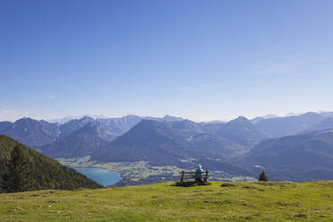 Rear view of senior male hiker sitting on bench while looking at Dachstein Mountains against blue sky - GWF06174