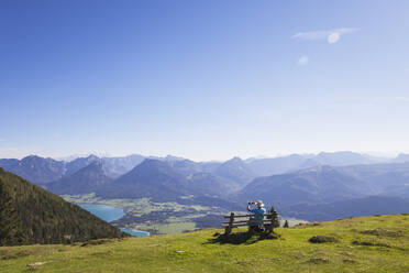Rear view of senior male hiker photographing while sitting on bench while looking at Dachstein Mountains against blue sky - GWF06175