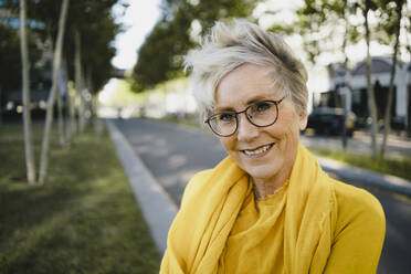 Porträt einer lächelnden reifen Frau mit grauen Haaren, die eine Brille und gelbe Kleidung trägt - KNSF06096