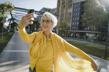 Portrait of smiling mature woman wearing yellow clothes taking selfie with smartphone - KNSF06102