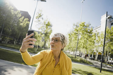 Portrait of mature woman wearing yellow clothes taking selfie with smartphone pouting mouth - KNSF06104