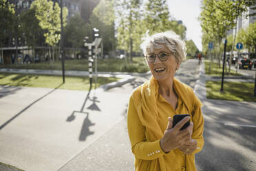 Portrait of happy mature woman with smartphone wearing yellow clothes outdoors - KNSF06108