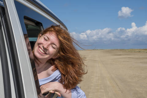 Unbekümmertes rothaariges Teenager-Mädchen mit Kopf aus dem Autofenster am Strand gegen den Himmel, lizenzfreies Stockfoto