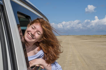 Unbekümmertes rothaariges Teenager-Mädchen mit Kopf aus dem Autofenster am Strand gegen den Himmel - LBF02623