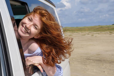 Porträt eines unbekümmerten rothaarigen Teenagers mit dem Kopf aus dem Autofenster am Strand gegen den Himmel - LBF02624