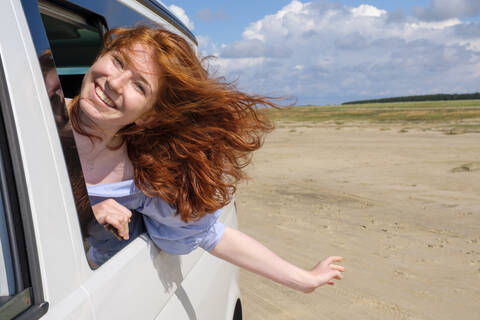 Porträt eines unbekümmerten rothaarigen Mädchens, das den Kopf aus dem Autofenster streckt und am Strand gegen den Himmel genießt, lizenzfreies Stockfoto