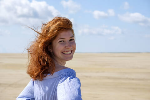 Lächelndes Teenager-Mädchen, das über die Schulter schaut, während es am Strand gegen den Himmel an einem sonnigen Tag steht, lizenzfreies Stockfoto
