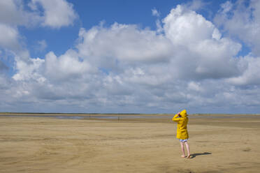 Voller Länge Rückansicht von Teenager-Mädchen trägt gelben Regenmantel beim Gehen auf Sand am Strand während des sonnigen Tages - LBF02634