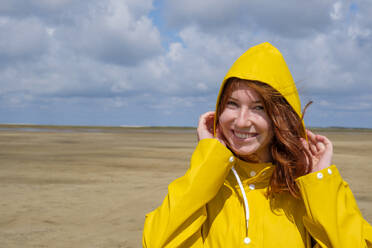 Close-up-Porträt von sorglos Rothaarige Teenager-Mädchen trägt gelben Regenmantel, während am Strand gegen den Himmel an einem sonnigen Tag stehen - LBF02635
