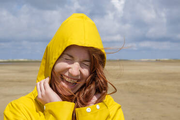 Close-up of carefree redhead teenage girl wearing yellow raincoat while standing at beach against sky on sunny day - LBF02637