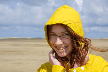 Close-up-Porträt von sorglos Rothaarige Teenager-Mädchen trägt gelben Regenmantel am Strand gegen den Himmel an einem sonnigen Tag - LBF02638