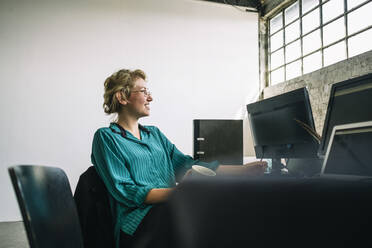 Nachdenkliche junge Geschäftsfrau, die lächelt und wegschaut, während sie am Schreibtisch in einem kreativen Büro sitzt - MASF13303