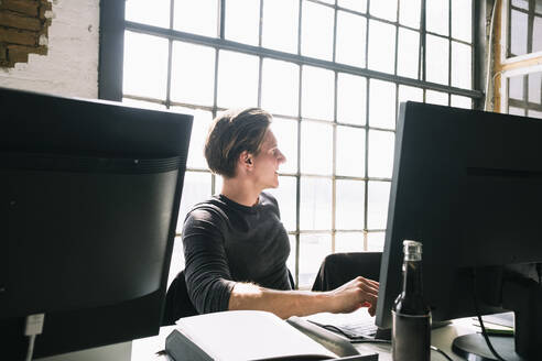 Junger männlicher Hacker, der wegschaut, während er an einem Computerschreibtisch an einem kreativen Arbeitsplatz sitzt - MASF13307