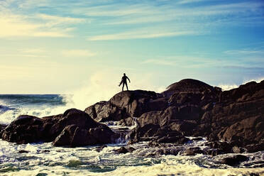 Silhouette eines männlichen Surfers, der die Wellen beobachtet, die an einem sonnigen Strand gegen die Felsen schlagen, Tofino, Kanada - FSIF04214