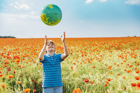 Glücklicher Junge, der einen Globus fängt, während er in einem Mohnfeld gegen den Himmel an einem sonnigen Tag steht, lizenzfreies Stockfoto