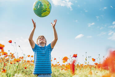 Junge, der einen Globus fängt, während er in einem Mohnfeld gegen den Himmel an einem sonnigen Tag steht - MJF02386