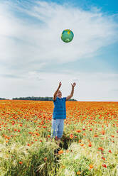 Boy catching globe while standing in poppy field against sky - MJF02396