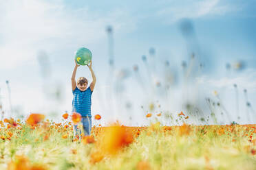 Happy boy holding globe while standing in poppy field against sky on sunny day - MJF02397