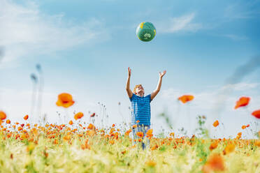 Boy catching globe while standing in poppy field against blue sky on sunny day - MJF02398