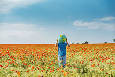 Pre-adolescent boy holding globe while standing in poppy field against blue sky - MJF02400