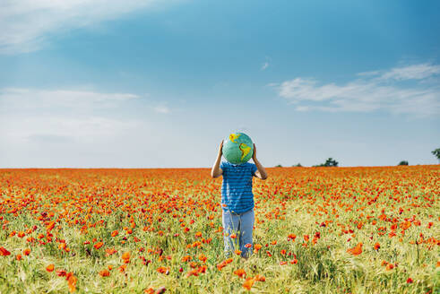 Ein Junge im Vorschulalter hält einen Globus, während er in einem Mohnfeld vor blauem Himmel steht - MJF02400
