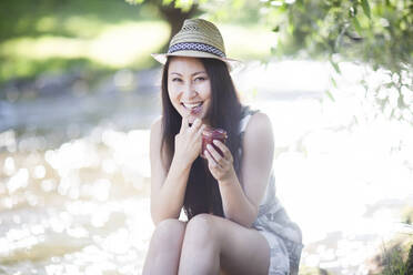 Smiling young woman wearing a straw hat, sitting at the riverside and nibbling - SGF02404