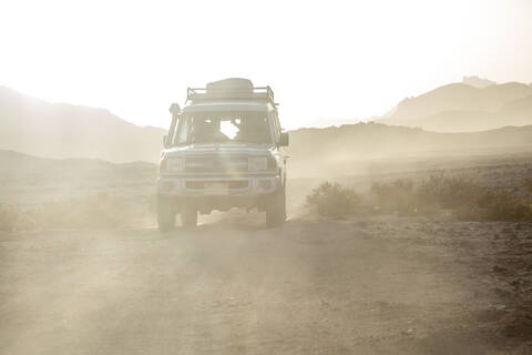 Geländewagen auf unbefestigter Straße inmitten von Staub in der Wüste gegen den Himmel bei Sonnenuntergang, lizenzfreies Stockfoto