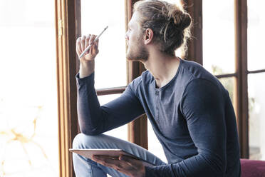 Nachdenklicher junger Mann zu Hause mit einem digitalen Tablet, der aus dem Fenster schaut - JSRF00443